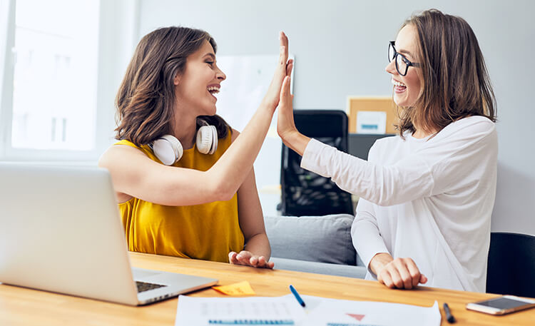 two women high-fiving while sitting at a table with a laptop